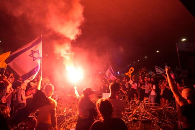 A crowd of people raise arms and yell, one holding an Israeli flag. A person near the centre holds a flare, casting a red light over everything.